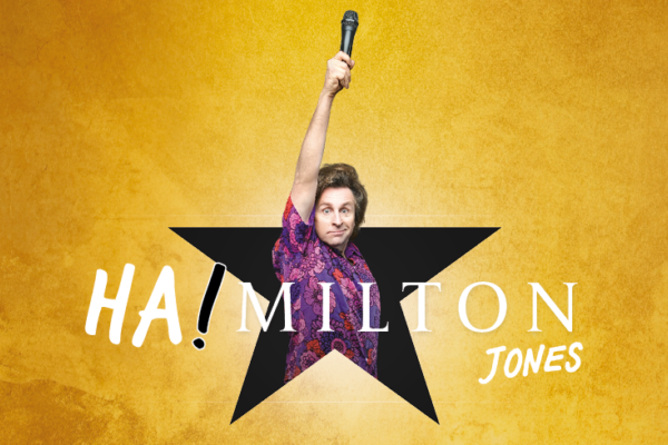 Milton Jones stands in a black star holding a microphone in the air with a gold backdrop