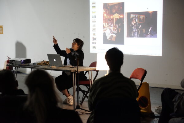 A person gives a lecture to an group of people and sits at a table with a laptop in front of them.  The person is holding a gesture with their hands and pointing to the ceiling and there is a projected image lighting up the wall behind them.