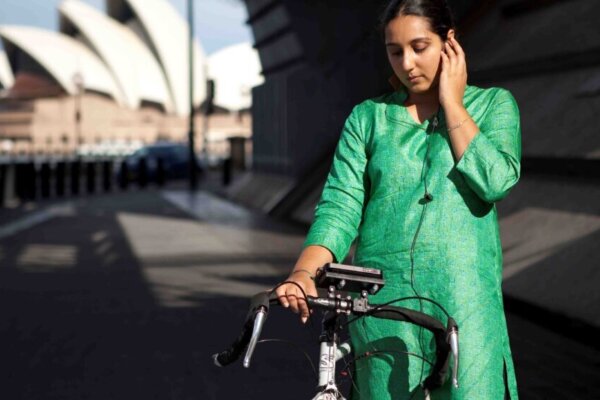 A woman in a green tunic stands under a wide bridge, astride her bicycle, holding an earpiece to her ear.