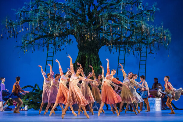 A large group of dancers all dressed in orange tones dance in front of a large tree in the center of the stage in the live screening of The Winter's Tale at Corn Exchange Newbury