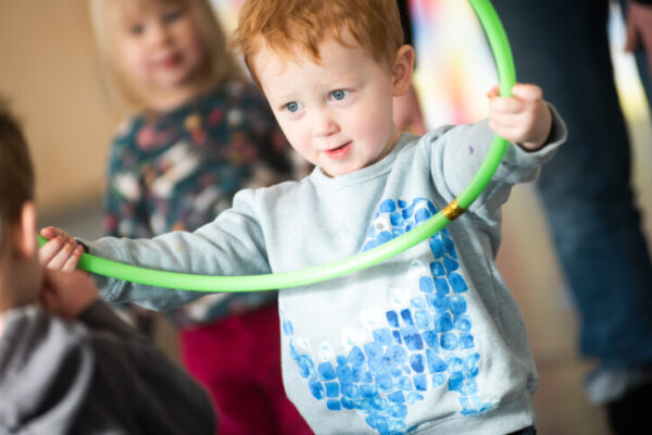 This is a photo of a ginger toddler holding a hula hoop. There are other children around him and the legs of an adult in the background.
