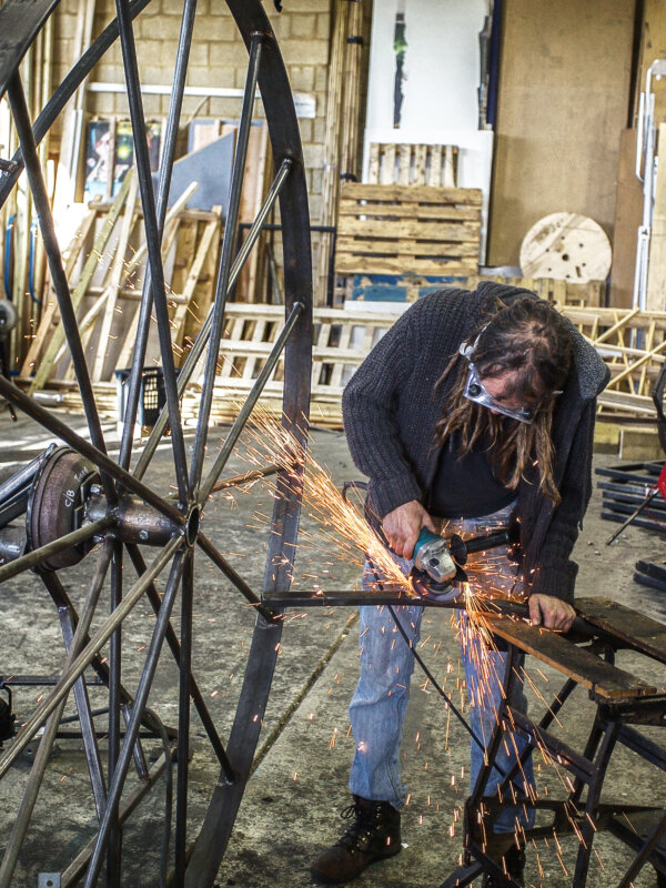 Image is taken in 101's metal/wood workshop. One person is captured fabricating a metal frame. To the left of the image is a large metal wheel with spokes. The person is using a hand tool and orange sparks are flying from the work piece.