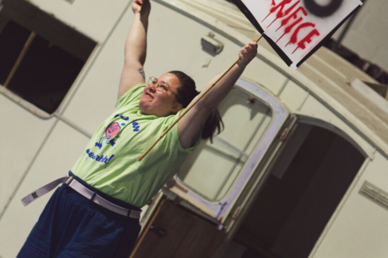 Artist standing outside an open caravan door, both arms are raised to the sky is a strong stance. In one hand they hold a protest sign that reads "NO SACRIFICE". the person is holding a passionate but hard expression on there face.