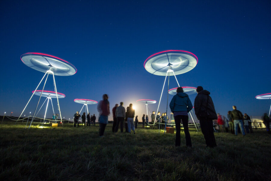 Image is of large spinning metal structures which are lit up against a dark blue sky. Some spectators gather to look up at the moving installations.