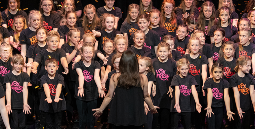 A photo of a woman facing towards a group of children singing and all performing a dance move in unison. They are all wearing black t-shirts which say VoxFresh on them.