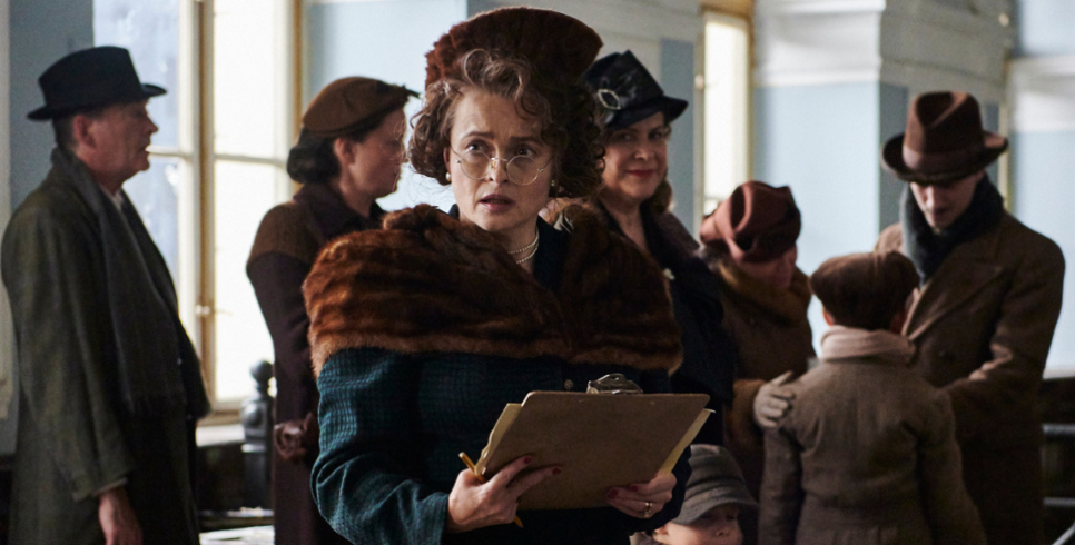 Helena Bonham Carter stands in a busy room wearing a fur scarf and hat with a clipboard in hand.