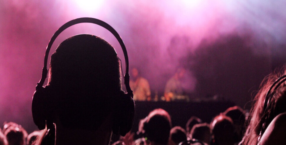 Against a pink, smokey background we see the back of a person's head. They have short hair and are wearing headphones.
