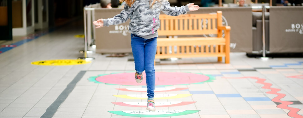 A young girl in a shopping centre, hopping along rainbow-coloured floor designs.