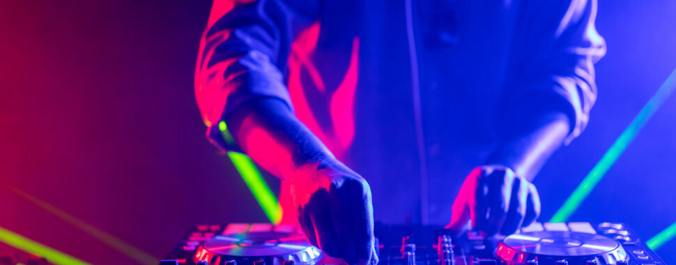 We see the hands of someone stood behind DJ decks, playing music. Coloured lights shine on them.