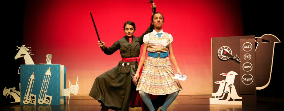 A girl on a red stage with paper cuts in the background as a woman holds her pony tail in the air