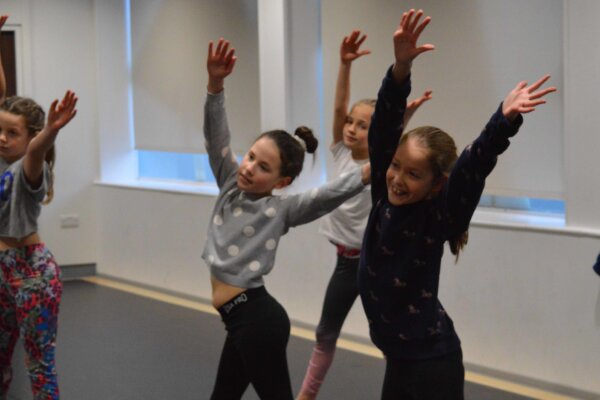 An action shot in a rehearsal room of young dancers with smiles on their face holding a pose with their arms in the arm pointing to one side.