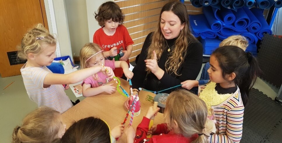 There is a table with children sat around playing with pencils. An adult is sat with them talking with them and leading them on the workshop.