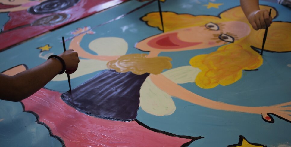A painting of a cartoon fairy with young people's hands holding paintbrushes and painting the fairy.