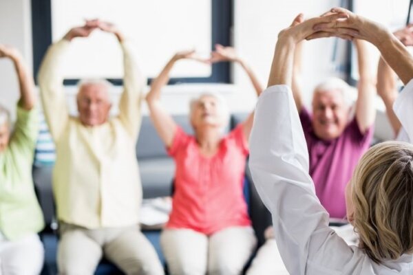 A circle of elderly people are smiling and stretching over their head facing an instructor.
