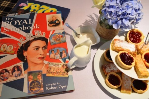 On the left hand side of a table, there is a book called The Royal Scrapbook with an image of the late Queen Elizabeth. Next to it, is a small jug of milk and to the right of tat is a two-tiered cake stand with a variety of cakes and sweet treats. Behind this is a blue floral plant.