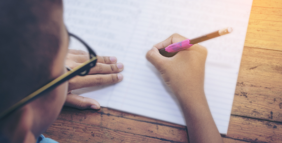 An image taken at birds eye perspective of a young boys head who is wearing glasses and is writing on piece of paper with a pencil.