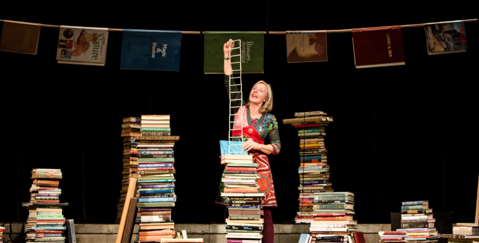 A woman is on stage with lots of tall piles of books around her. She is holding a mini ladder on top of one of the piles of books. There is also some bunting behind her.