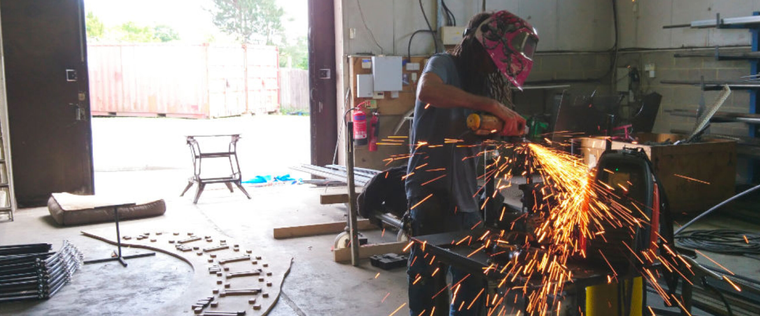 Image taken in 101 Metal workshop. A person wearing a welders visor fabricates a metal frame. Bright orange sparks are flying from the hand held tool.