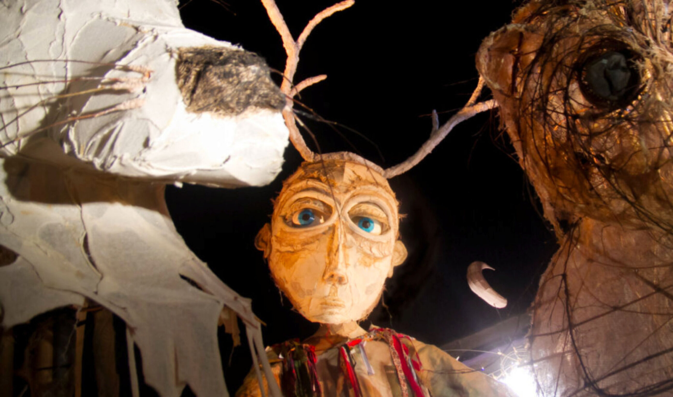 An ominous giant puppet face looking into camera with large blue eyes. 2 large animal puppets stand along side it. Image has black skies in background.