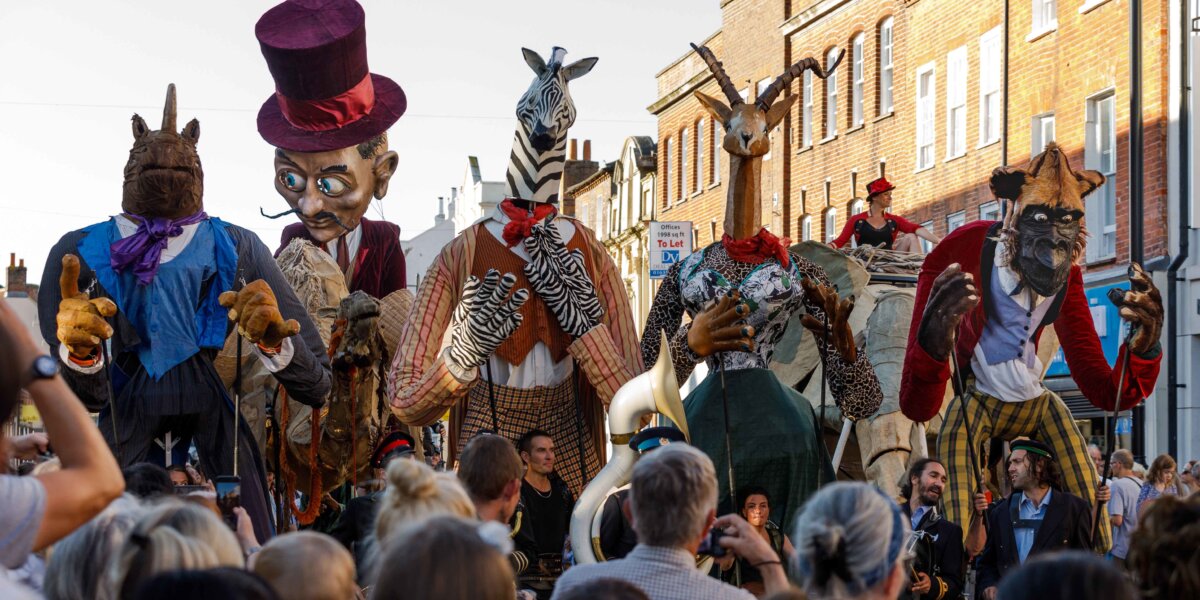 Five giant puppets walk side by side down the street. From left to right, a Rhino in a blue coat, a man with a black moustache and a burgundy top hat, a zebra in a tweed suit, an antelope in a corset, a monkey in a red jacket and yellow chequered trousers. Are all double the height of the people in the audience.