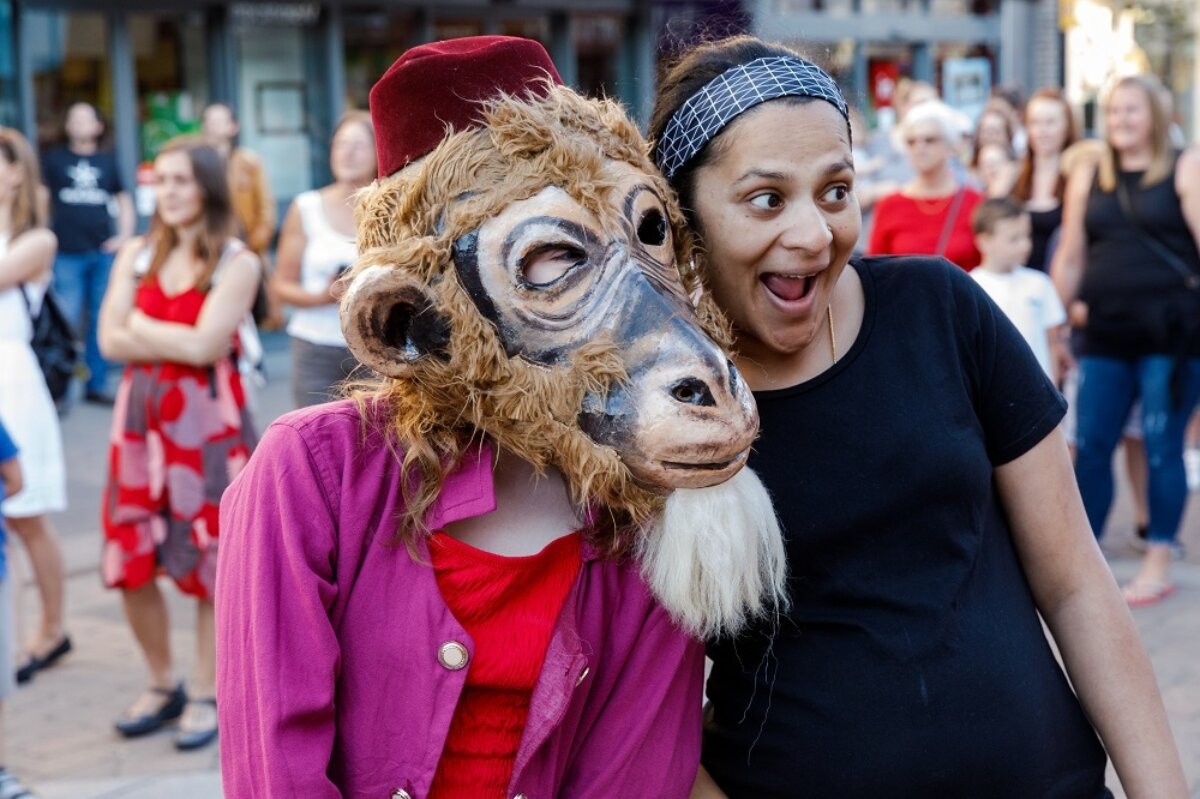 A woman in a suit with a giant monkey mask and a top hat poses for a photo with a smiling woman in a black t-shirt and a black and white bandana.