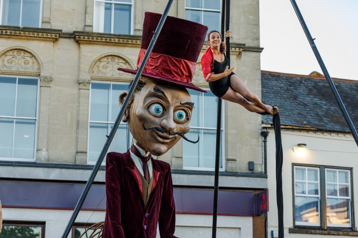 A giant puppet man with a black pencil moustache and a burgundy top hat stands in Newbury Market Place looking at a female acrobat suspended in the air from a tripod. The woman looks to be the size of the puppet's head and tall hat.