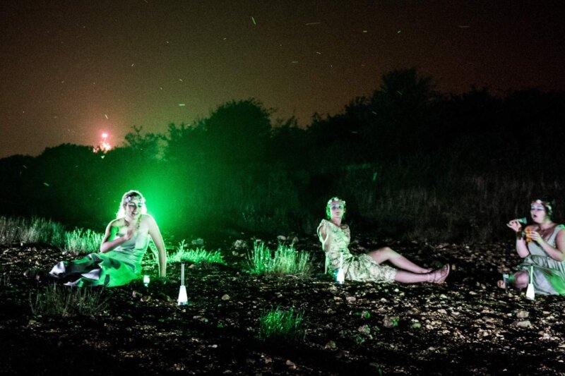 In a field in the dark, three women sit on the grass in pale dresses lit up with a green glowing light. It looks etherial.