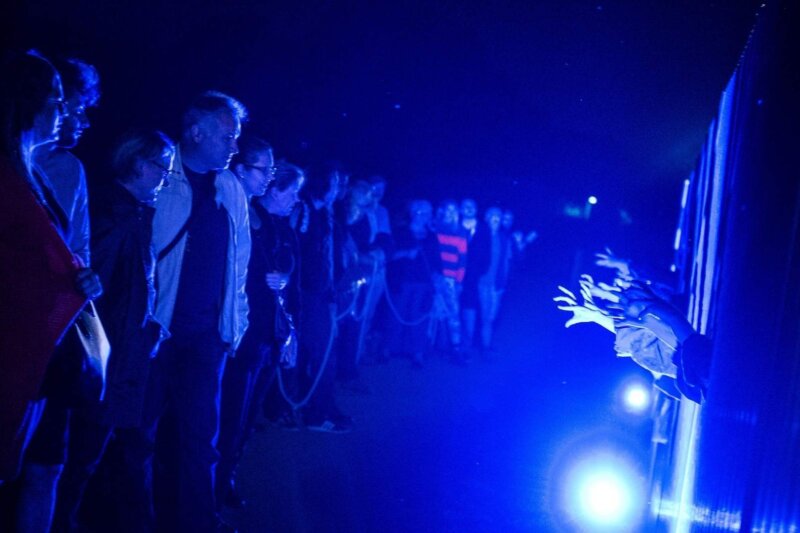 An audience stand in the dark in front of a glowing blue cage structure that has dancers hands reaching out of it.