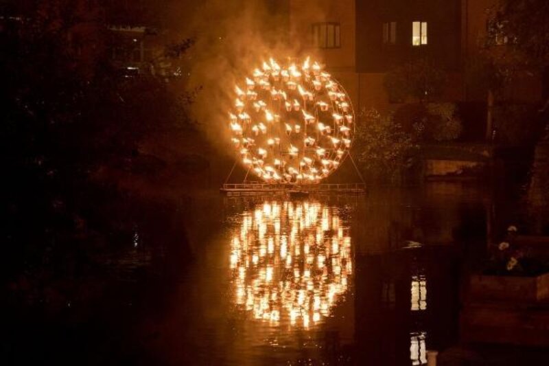 A sphere of metal arms hovers above a canal in the dark. The arms have a cup filled with fire on each end making the sphere glow orange. The reflection on the still water below is perfect.