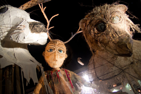 Against a dark sky, a wicker and paper giant puppet man looks down at the camera, next to the head of a giant bear made from similar natural materials.