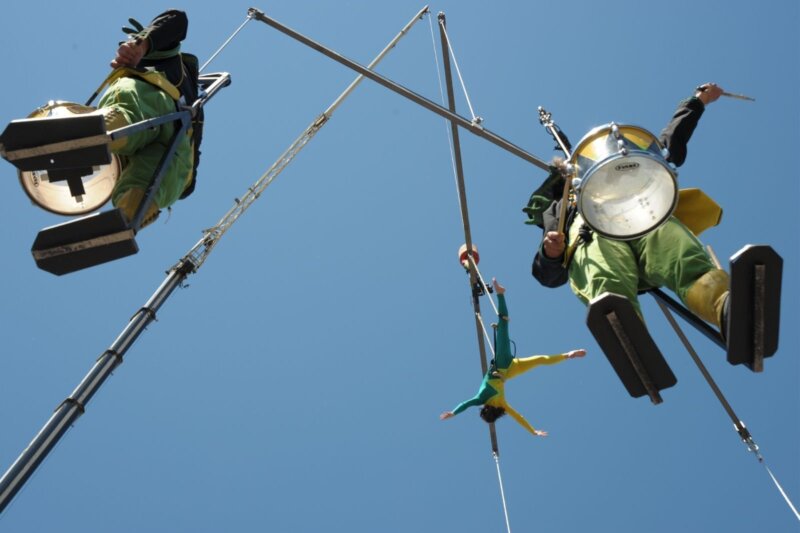 This photo is taken from below looking up to the very blue sky. It features three people attached to a large wooden aerial mobile suspended in the air. Two of the people are dressed in green and yellow toy soldier costumes playing the drums, one is in dressed in a green and yellow Lycra outfit performing aerial acrobatics.