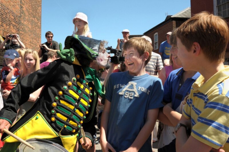 A man dressed in a green and yellow little drummer boy costume with white paint on his face leans towards a boy in a crowd of people pulling a silly face, puckered up as if about to kiss the boy. The boy is wearing a blue t-shirt and laughing at the man.