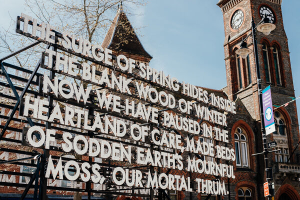 A closeup of a sign made up of lights with the words 'The surge of spring hides inside the blank wood of winter now we have paused in the
heartland of care, made beds of sodden earth’s mournful moss, our mortal thrum'. The backdrop is Newbury Town Hall.