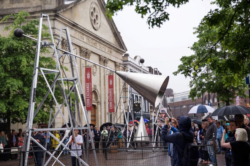 A large metal bell structure is swinging back and forth in front of the Corn Exchange building Newbury. The Market place is rainy and audiences stand in coats and under umbrellas.