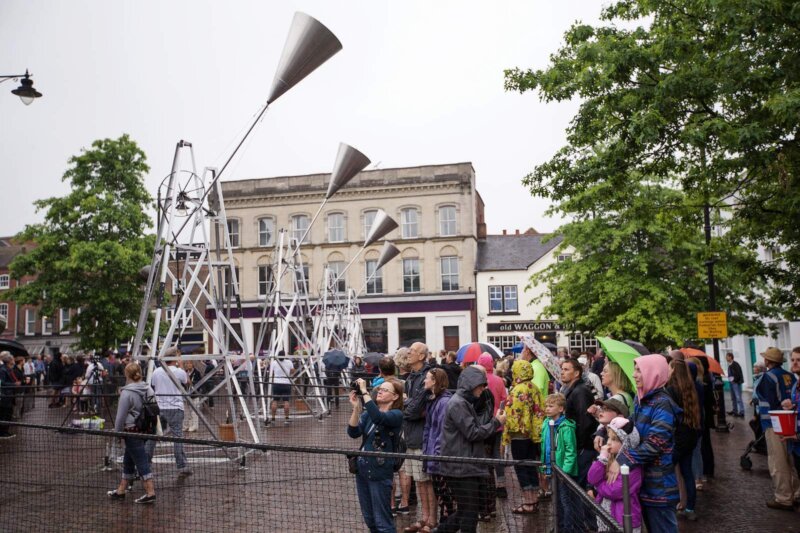 A line of large metal bell structures all swung up towards the sky sit in Newbury Market Place.