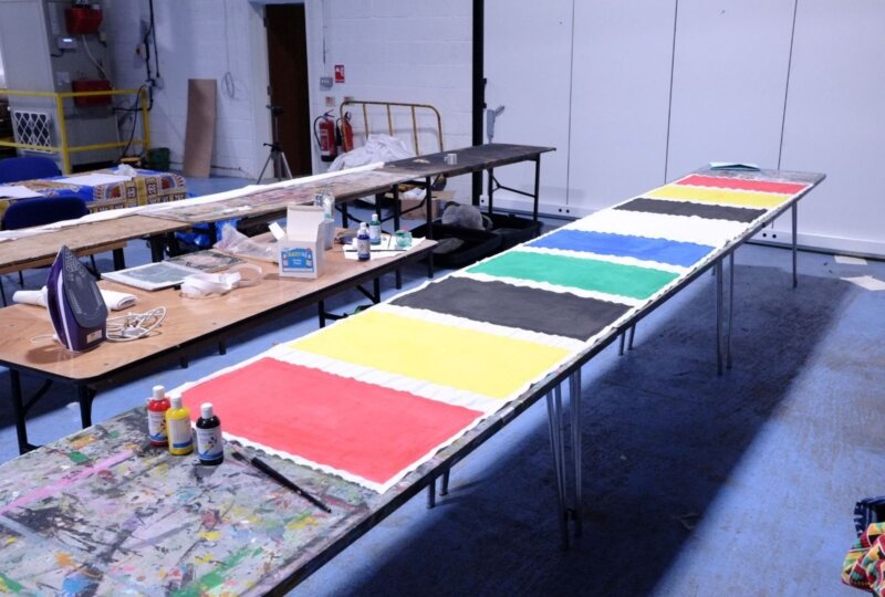 Two long wooden tables, one with a selection of craft items on, and one with a long strip of fabric painted in strips of red, yellow, black, blue and green.
