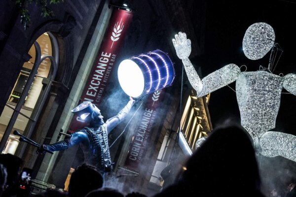 Outside the Newbury Corn Exchange in the dark, a giant puppet made of light is high above the heads of the crowd as well as a drummer in a costume illuminated with white lights.