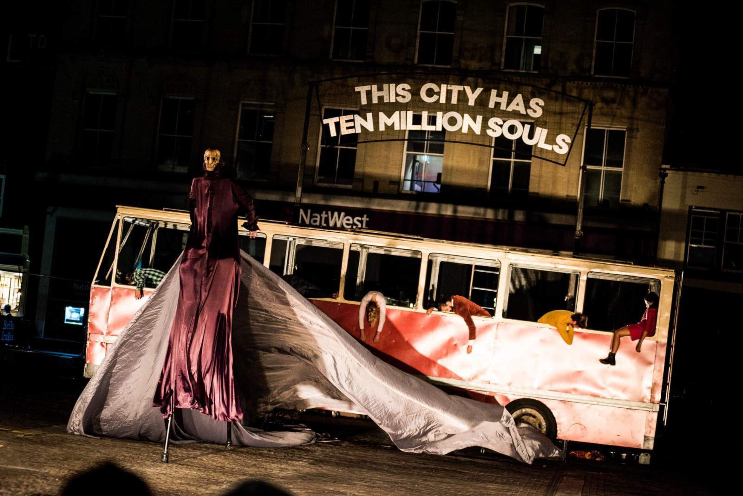 A scary scene, a giant man on stilts with an expressionless mask over the face, wearing a red hood that flows for meters behind him, stands in front of an old red bus. People hang like rag dolls from the bus windows, and above the roof a silver sign reads 'this city has ten millions souls'.