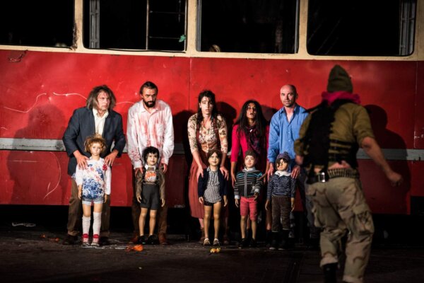 Five adults stand in a line in front of an old red bus, each of them holding a child doll infant of them. A man in khaki combat gear runs towards them.