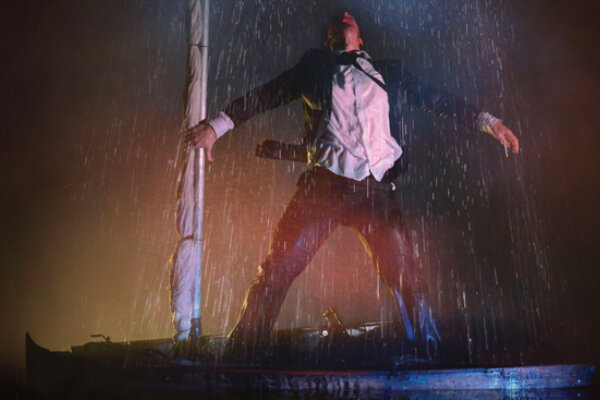 A man in a black suit and white shirt stands in a boat, head thrown back, one hand gripping the boat's mast as a fake raincloud rains water down on him.