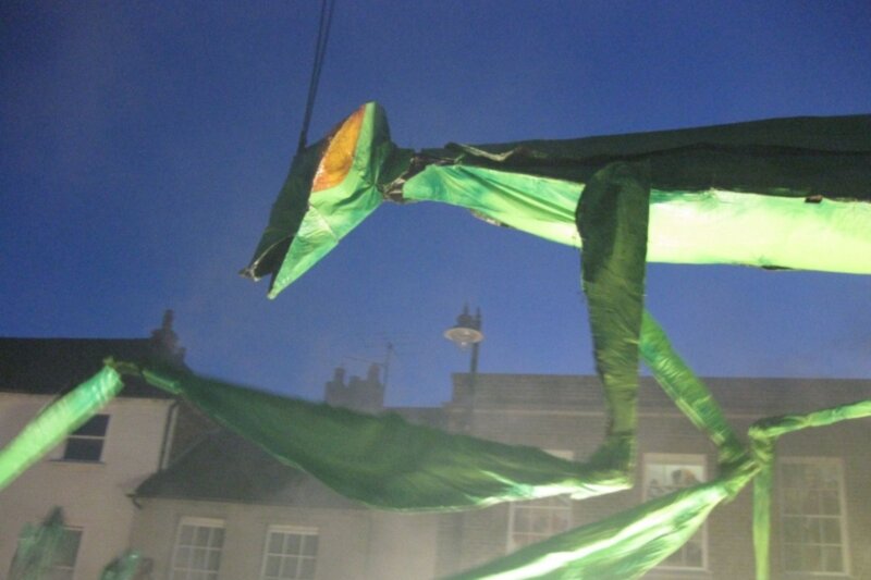 A large, bright green praying mantis puppet next to buildings in Newbury.