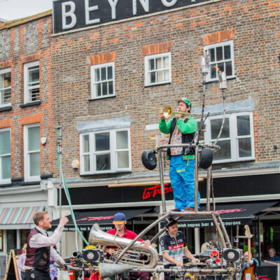 A man stands on a scaffold playing a brass instrument in a high street.