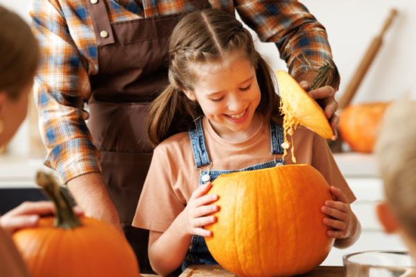 A photo of a little girl smiling while she carves a pumpkin with the guidance of an adult behind her. You can also see blurred out and facing the other way a woman and a young boy.
