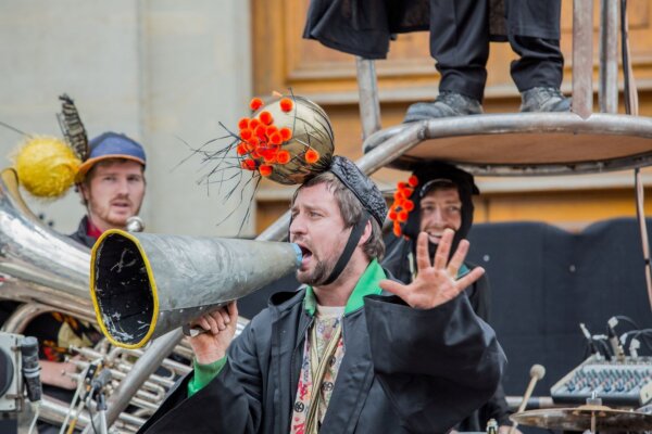 Image of 4 artists performing a live outdoor show in Newbury. The artists are dressed in a mix mash of baggy casual clothes inspired by brass band uniform. The artists in the background are playing instruments and the artist in the center is talking into a prop megaphone with their other hand stretched out in a 'stop' gesture.
