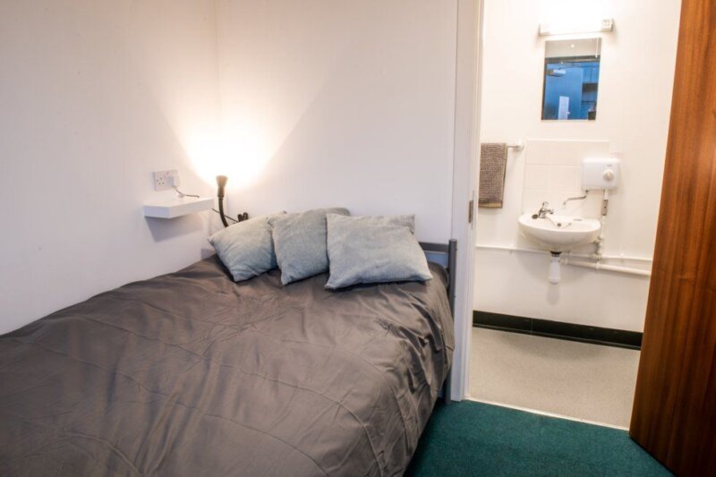 An interior of one of 101's portacabins, showing a double bed with grey bedding and white pillows. The open doorway is visible to an ensuite bathroom.