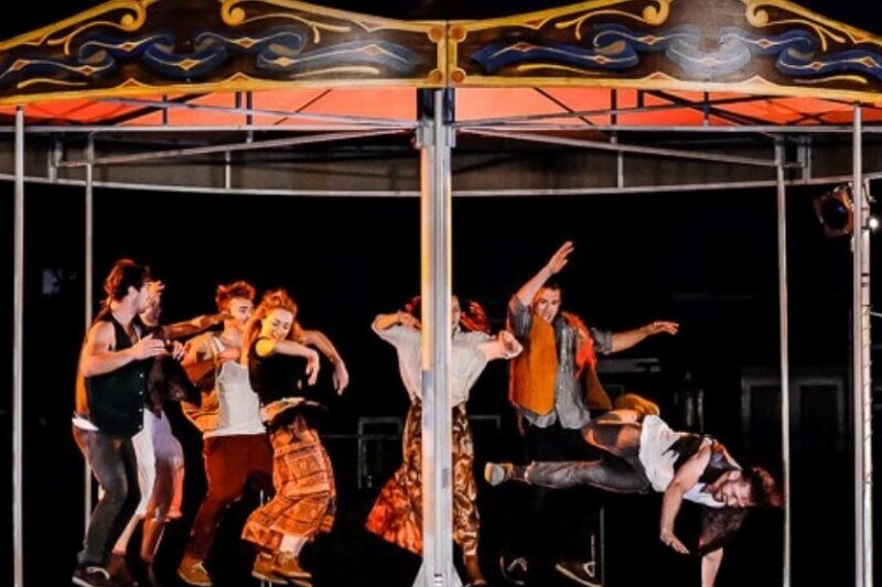 6 men and women dance jubilantly on a carousel - one goes down on one hand with his legs off the ground.