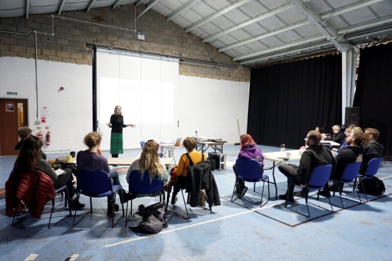 A woman speaking in front of a projection screen and listeners sat in a row of chairs with their backs to the photographer.