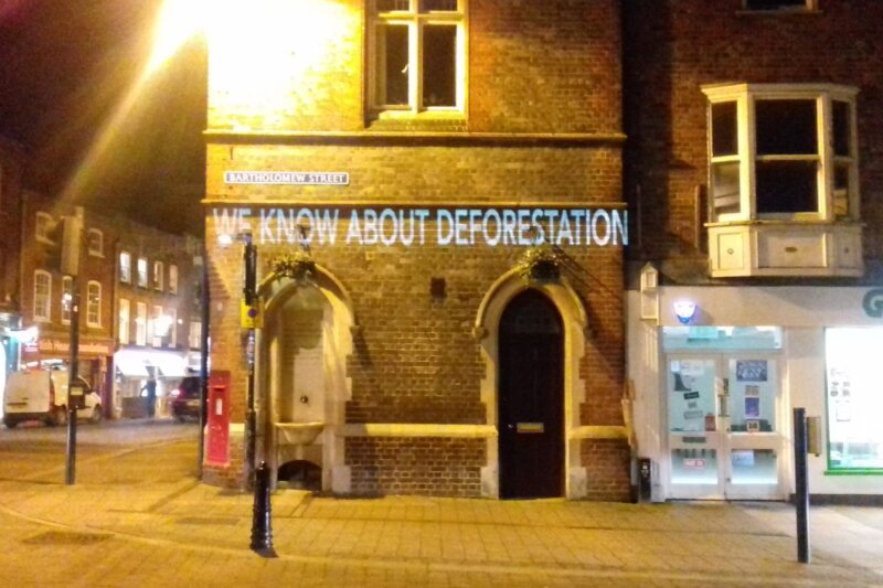 White words projected onto a brick wall at night in Newbury town centre. The words read:
WE KNOW ABOUT DEFORESTATION