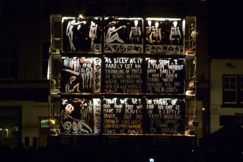 A giant canvas on a scaffold with white letters lit up against a black background and painted images too.