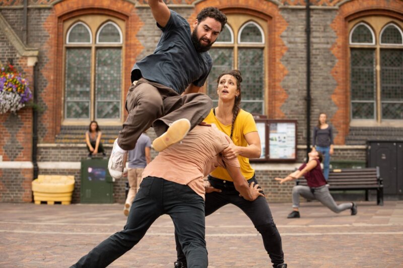 One dancer pushes another back, while a third vaults over the first dancer's back. The dancers are in Newbury market place and wearing everyday clothes, coloured jeans and t-shirts.
